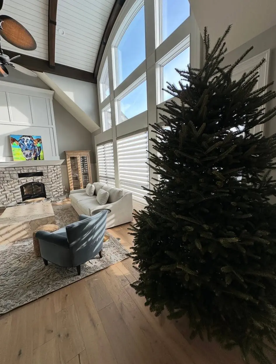A tall fresh-cut Christmas tree set in a two-story living room with a stone fireplace